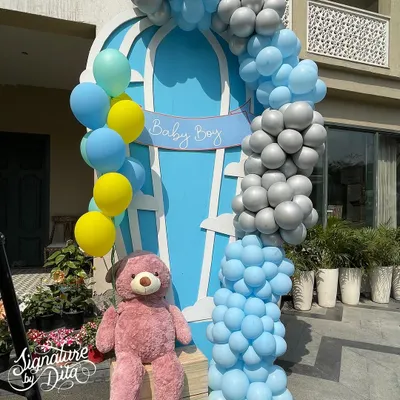 A beautiful 'Baby Boy' welcome setup for a baby shower, featuring a hot air balloon motif, a cascade of blue, yellow, and silver balloons, and a cuddly teddy bear.