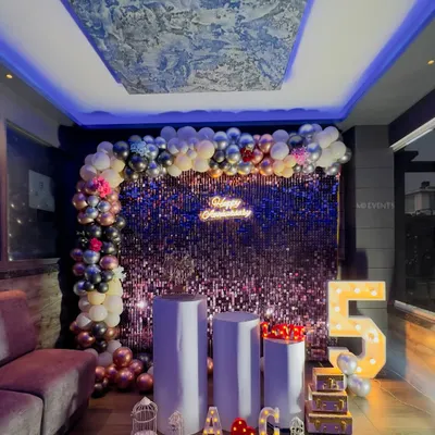 A glamorous 5th-anniversary party setup in a lounge. I used a purple shimmer wall, a multi-colored balloon garland, and large marquee numbers to create a vibrant and celebratory atmosphere.