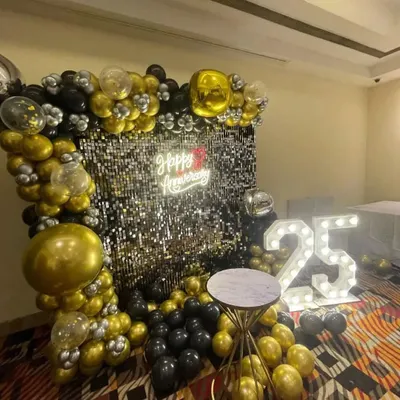 A stunning 25th-anniversary decoration in black, gold, and silver. This setup includes a balloon arch, a silver sequin wall, a "Happy Anniversary" neon sign, and large marquee numbers.