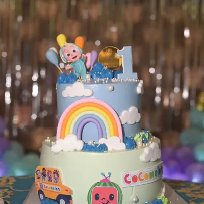 A colorful two-tier Cocomelon cake for a first birthday. This design features JJ, the iconic school bus, and a bright rainbow, creating a cheerful centerpiece for the little one's big day.
