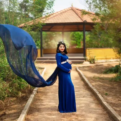 An expecting mother stands on a park pathway in a flowing royal blue maternity gown, the fabric caught in the wind to create a dramatic, wing-like effect. This outdoor maternity portrait captures a moment of power and beauty against a natural, sunlit backdrop.