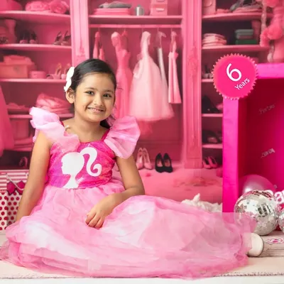 A six-year-old girl smiles brightly in her Barbie-themed shoot, sitting beside a life-sized Barbie box in a room filled with pink.