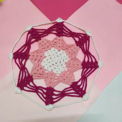 This is the Karamella doily, an intricate design featuring layers of stitches in shades of pink and white. It can be used to decorate a table or hung as a beautiful piece of wall art.