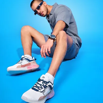A modern shoe campaign featuring a model in a casual pose against a vibrant blue background. The wide-angle perspective emphasizes the stylish sneakers, making them the focal point of this commercial shot.