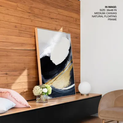 This abstract painting features a dramatic sweep of white and black with a touch of gold, creating a sense of movement and elegance. Displayed in a natural floating frame, it adds a sophisticated, contemporary touch to a modern console table.
