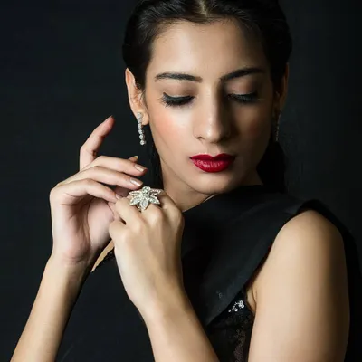 An elegant low-key portrait of a woman in a black saree. The lighting gently illuminates her face and hands, highlighting her red lipstick and diamond jewelry against the dark background.