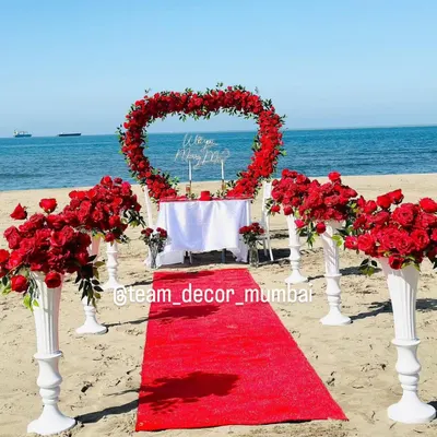 A full view of the romantic beach proposal setup I offer in Mumbai. It includes a red carpet aisle, a heart-shaped arch of red roses, floral pedestals, and a beautifully set table for two, right by the sea.