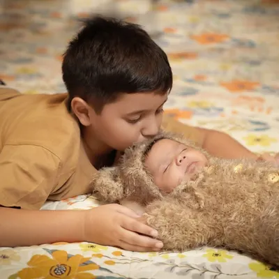 A moment of pure tenderness between brothers. An older brother lies on a floral patterned floor and gives a gentle kiss to the forehead of his baby sibling, who is sleeping peacefully in a fuzzy bear costume.