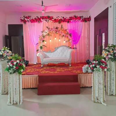 An elegant stage setup for a reception or engagement, featuring a white chaise lounge against a backdrop of pink curtains. The look is completed with a floral arch and matching flower-wrapped pillars.
