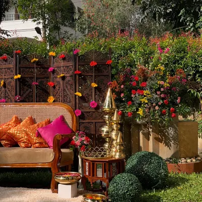 A beautiful outdoor seating area for a Mehandi function. The setup features a traditional carved wooden screen, vibrant pink and orange cushions, and brass decor, creating a rich and colorful ambiance.