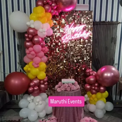 A glamorous birthday setup featuring a gold shimmer wall, a 'Happy Birthday' neon sign, and a colorful balloon arch in shades of pink, yellow, and white.