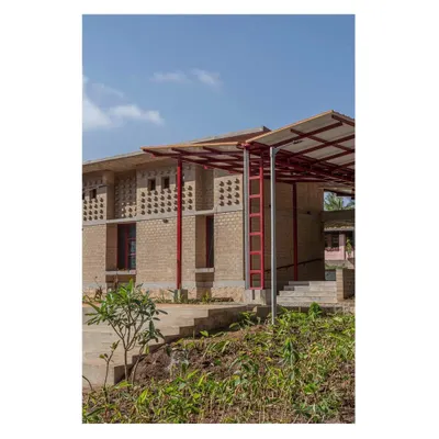 An exterior view showcasing the building's form, which integrates a central courtyard and uses perforated brickwork for natural cooling. The red steel structure provides a modern contrast to the traditional earth construction.