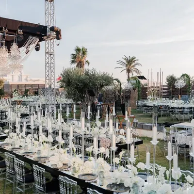 An outdoor reception with long tables adorned with a sea of candles and white florals. The modern, transparent chairs and dramatic chandeliers create a stunning contrast against the open-air setting, crafting an unforgettable dining experience.