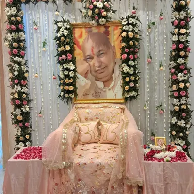 A classic and complete altar arrangement for a Guru Ji satsang. The setup includes floral pillars, a decorated throne with custom pillows, and side tables for offerings, all set against a clean white curtain backdrop.