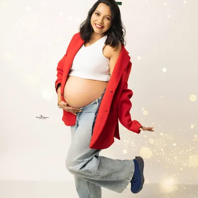 A fun and festive Christmas-themed solo portrait. The mother-to-be is dressed in a cheerful red blazer and jeans, kicking up her heel with joy.