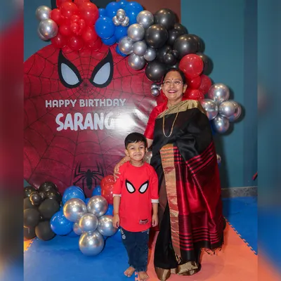 The birthday boy, dressed in his Spiderman shirt, stands proudly with his grandmother in front of the custom superhero backdrop, a perfect photo opportunity.