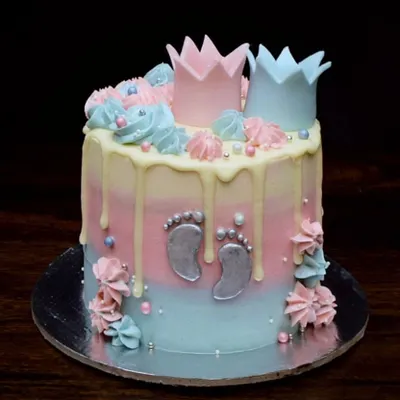 A sweet baby shower cake perfect for a gender reveal party. It features a pink and blue ombre design, a white chocolate drip, and is topped with little pink and blue fondant crowns. The tiny silver baby footprints are my favorite part.
