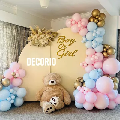 A classic "Boy or Girl" gender reveal party setup. This design uses a neutral backdrop with gold lettering, a large teddy bear, and a mix of pink and blue balloons to build anticipation for the big reveal.