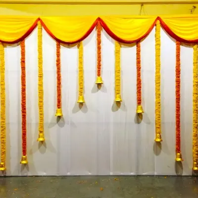 A simple and traditional Haldi ceremony backdrop for home. The decor uses yellow and orange marigold garlands with temple bells hanging from a yellow fabric drape.