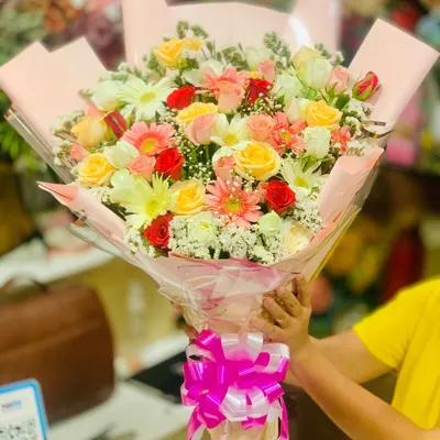 A large, vibrant bouquet filled with a mix of fresh flowers. This arrangement includes yellow roses, red spray roses, white daisies, and pink gerberas, all wrapped in pink paper with a matching bow.