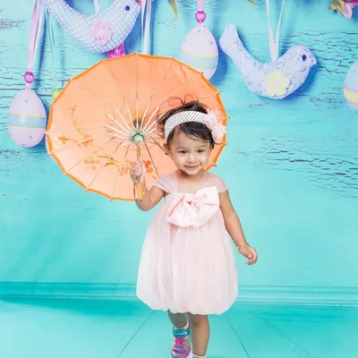 A sweet toddler in a pink dress holding a bright orange umbrella. The colorful props and cheerful blue background create a very happy and playful portrait.