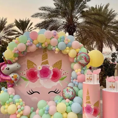 A beautiful outdoor unicorn party setup with a circular backdrop featuring a unicorn face, surrounded by a delicate pastel balloon garland. Themed plinths are ready for the cake and treats.
