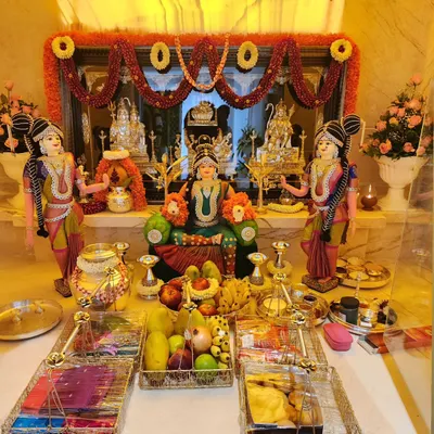 A complete Varamahalakshmi pooja setup with the goddess seated on a throne, surrounded by fruits, flowers, and traditional offerings. The decor includes a beautiful flower *thorana* in the background.