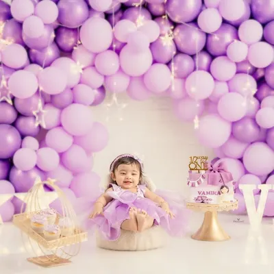 A perfect first birthday portrait. The birthday girl sits proudly in her purple dress, surrounded by a stunning balloon arch, a custom cake, and light-up letters spelling her initials.