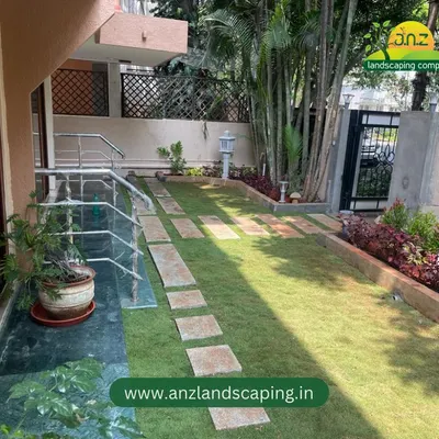 A complete front yard makeover in Indiranagar, featuring a new Mexican grass lawn bordered by colorful plants and a functional stone pathway leading to the entrance.