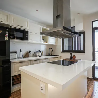 A modern kitchen with off-white shaker cabinets, a quartz island with a built-in cooktop, and a striking stainless steel chimney.