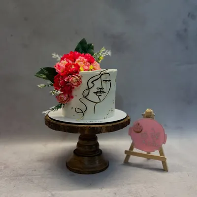This cake features a minimalist one-line art face, hand-piped onto smooth white buttercream. The design is complemented by a vibrant bouquet of fresh flowers.