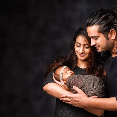 A classic newborn portrait with parents. The dark, moody background creates an intimate atmosphere, drawing attention to the parents' gentle hold and loving gaze upon their new baby.