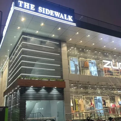 A night view of the completed commercial building, 'The Sidewalk'. The facade features horizontal louvers with integrated LED lighting, creating a clean, modern, and inviting look for the retail spaces within.