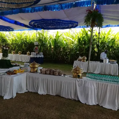 A beautiful outdoor catering setup under a tent for a day event. The multi-level buffet arrangement allows for a grand display of food against a lush green backdrop, perfect for weddings, receptions, and society events.