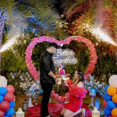 The pinnacle of a surprise proposal. The moment of the question, complete with fireworks, a heart-shaped floral arch, and a path of rose petals.