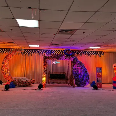 A beautiful stage setup for a baby shower featuring a floral moon-gate, a decorated Sankheda jhula, and a peacock made of flowers. The warm lighting creates a welcoming atmosphere for the celebration.