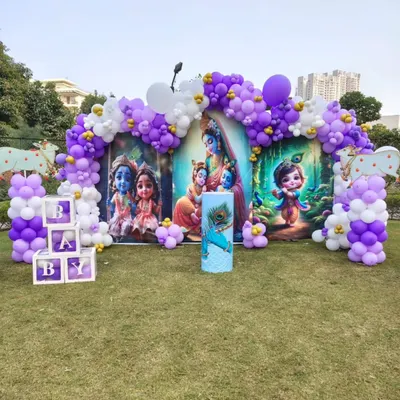 A unique Krishna-themed baby shower. This outdoor setup features a custom backdrop with adorable illustrations of baby Krishna, framed by a purple and white balloon arch.