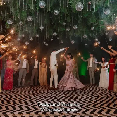 The couple makes their grand entrance, framed by a sparkling arch of light held by their loved ones. This is a classic Sangeet shot, full of magic and energy, set against a beautifully designed outdoor dance floor.