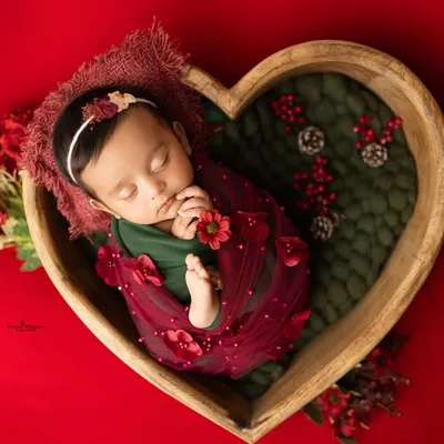 A wide shot of our Christmas-themed setup, with a peacefully sleeping newborn nestled in a heart-shaped bowl. The rich red backdrop and festive details create a warm, celebratory feel for your baby's first holiday photos.