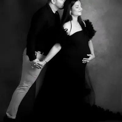 A full-length black and white portrait that feels like a classic dance pose. The contrast and lighting beautifully highlight the couple's connection and the elegant lines of the maternity gown.