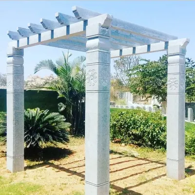 A beautifully crafted stone pergola with carved details on the pillars. This structure is set in a garden and provides a perfect frame for the surrounding plants.