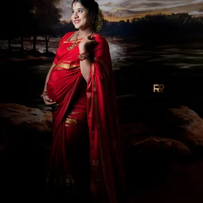 A portrait of timeless grace. The mom-to-be is dressed in a vibrant red saree, standing against a painted backdrop that evokes a serene, moonlit landscape. This is one of our signature traditional looks.
