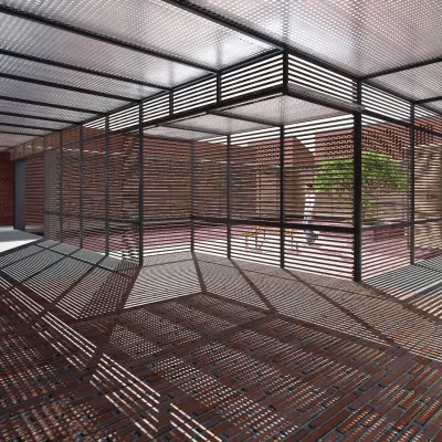 At Studio Tarai in Rudrapur, a louvered steel canopy casts a complex grid of shadows on the brick floor, creating a dynamic, patterned space that changes with the movement of the sun.