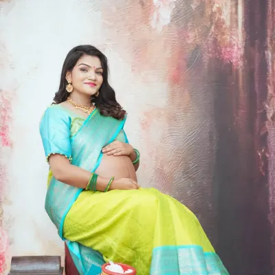 A graceful solo portrait of an expecting mother in a traditional silk saree. The painterly backdrop and classic props like decorated pots add a rich, cultural feel to this maternity photo.