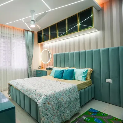 The final look of a beautiful kid's bedroom. The channel-tufted upholstered bed in teal is the star, complemented by striped wallpaper, overhead cabinets with gold trim, and playful geometric ceiling lights.