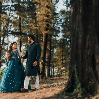 A couple posing in a beautiful forest setting. He wears a classic dark sherwani that perfectly complements her teal lehenga, showcasing my coordinated rental options.