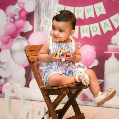 A perfect first birthday portrait using our pink party backdrop. The setup includes a "Happy Birthday" banner, balloon decorations, and a wooden chair, creating a complete party scene for your celebration.