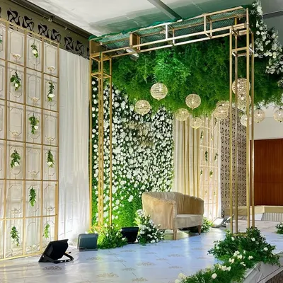 A wide view of this spectacular reception stage. The design features a lush green floral ceiling, a white flower wall, and elegant gold-toned structural elements for a modern and glamorous look.