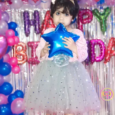 A young birthday girl dressed as a princess, posing in front of her colorful balloon and silver foil curtain backdrop.
