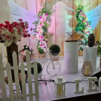 A whimsical and romantic backdrop featuring angel wings and fresh floral arches. The rustic wood-paneled background and white picket fence add to the garden-party charm.
